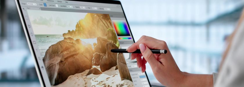 Closeup of a photo editor's hand using a stylus to edit a scenic beach photo on a touchscreen.