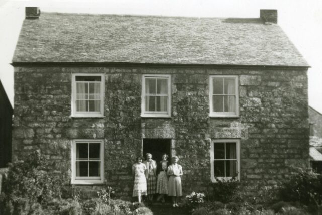 Original black-and-white photo of a stone house with four women standing in the doorway, showing a soft, slightly blurred vintage image before AI restoration.