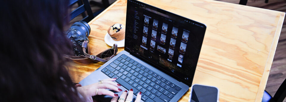 Photographer browsing a photo library on a laptop next to a camera and lens at a cafe table