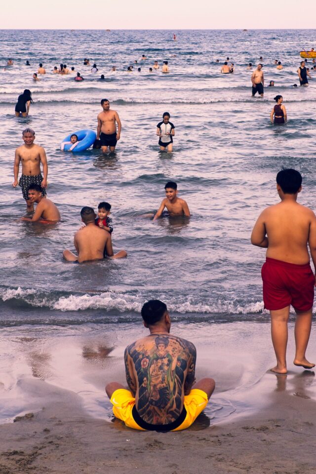 A late-afternoon beach scene from Da Nang, Vietnam — a solitary man watching the water while beachgoers fill the frame behind him. Finished in ON1 with a Warm vintage filter, color grading adjustments, Sunshine at 80% strength, and curve tweaks to arrive at the final look.