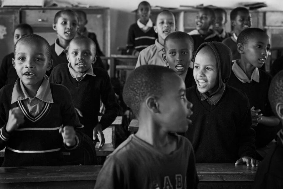 Black and white candid photo of a classroom of laughing, singing students in Tanzania