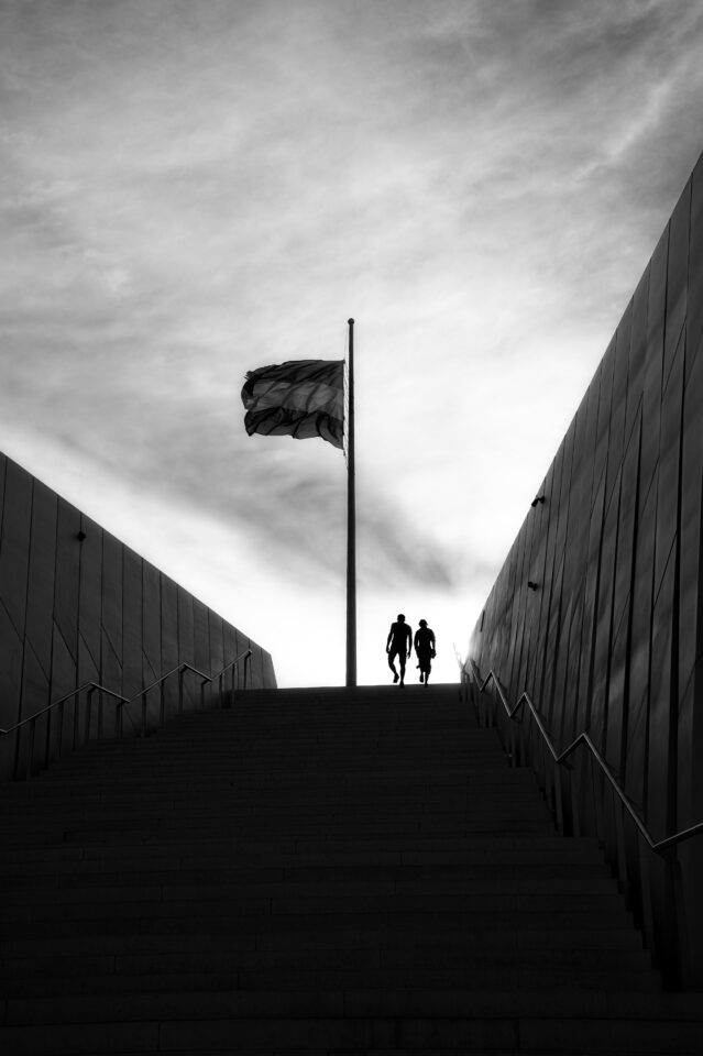 Two silhouetted figures walking up a dramatic staircase between converging geometric walls, black and white, with a flag overhead