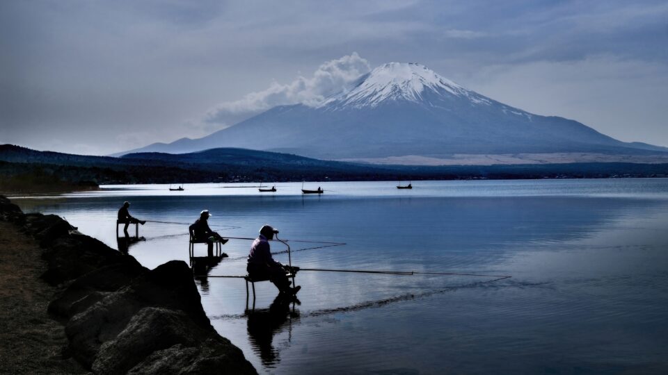 Three fishermen in silhouette on a rocky shore fishing on a still lake at dawn, with Mount Fuji in the background