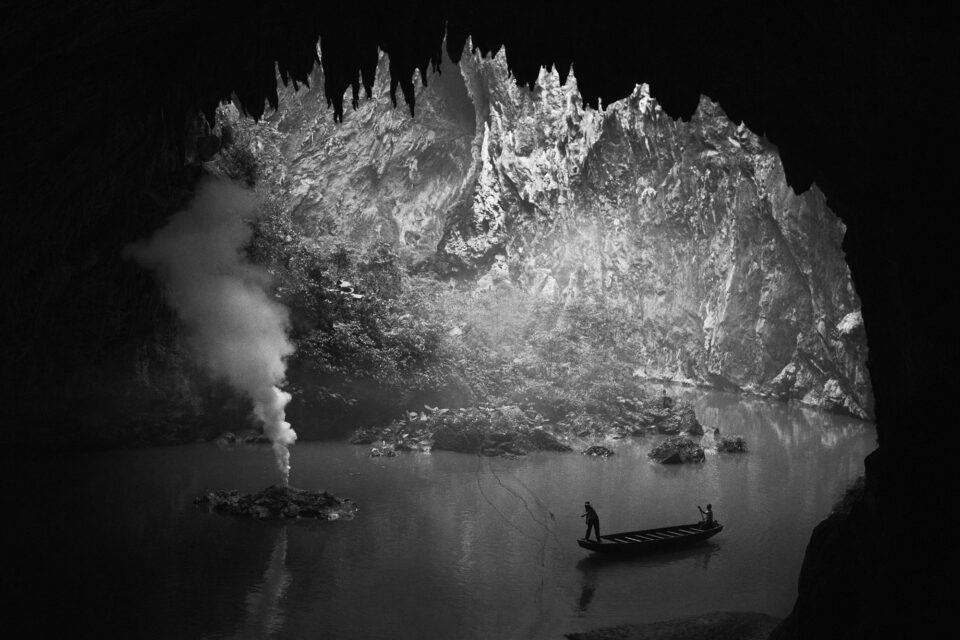 A small fishing boat with two fishermen inside a massive cave, with mist rising from the water, in black and white