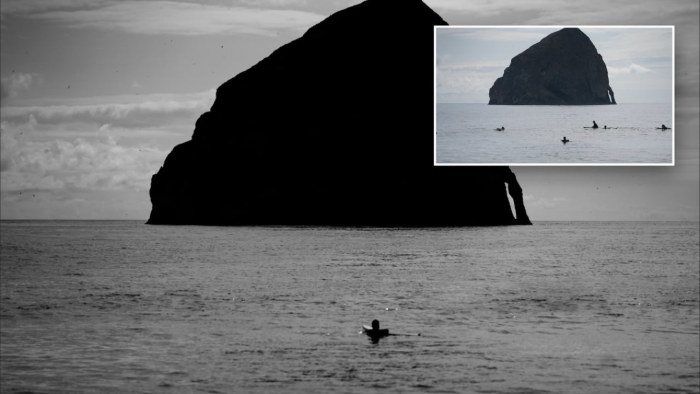 Black and white photo of a large sea stack and a single surfer, with an inset of the original frame showing several extra surfers in the water